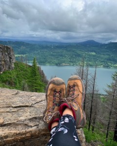 Landscape shot of the columbia river gorge with trees and the river and a pair of legs with hiking boots lounging on top of a rock. 
